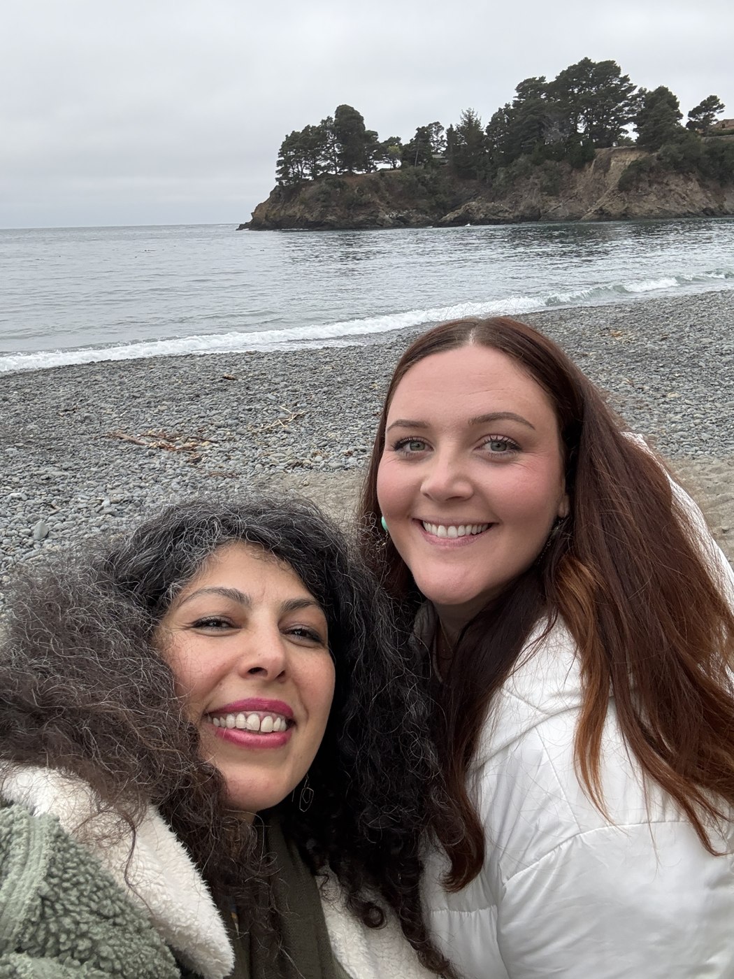 Armineh and Alexandra smiling together on a pebble beach by the ocean